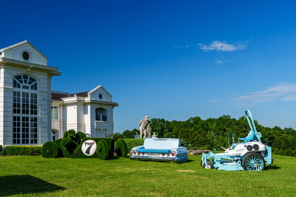 Blue and white custom lawn mower with flatbed and large spoked wheels on grass lawn near white building and statue