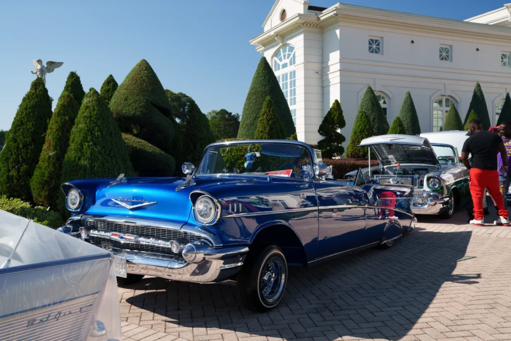 Blue Chevrolet Bel Air convertible and another two-tone Chevrolet with hood open at outdoor car event near white building and