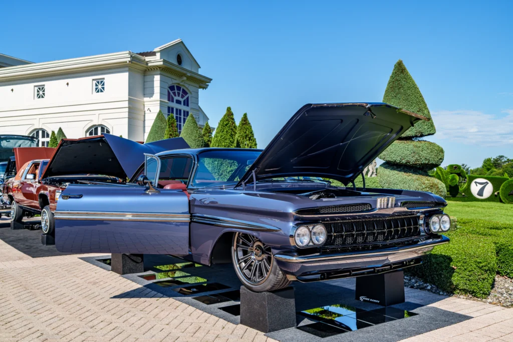 Blue 1959 Pontiac Bonneville with open hood and door at an outdoor car show with manicured bushes and a building in the backg