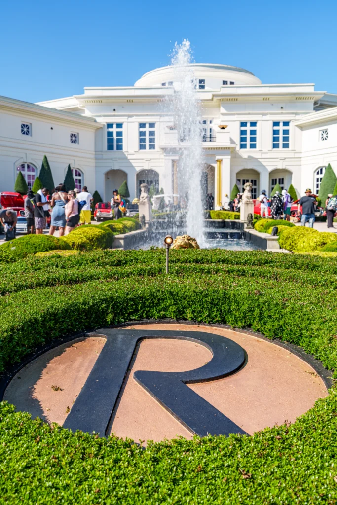 Fountain in front of a large white building with people and classic cars gathered around it on a sunny day