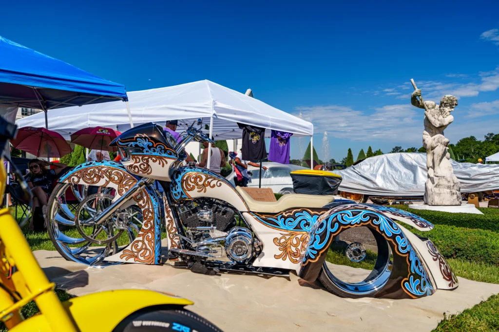 Custom chopper motorcycle with intricate blue and brown scrollwork paint parked near tents and a statue outdoors