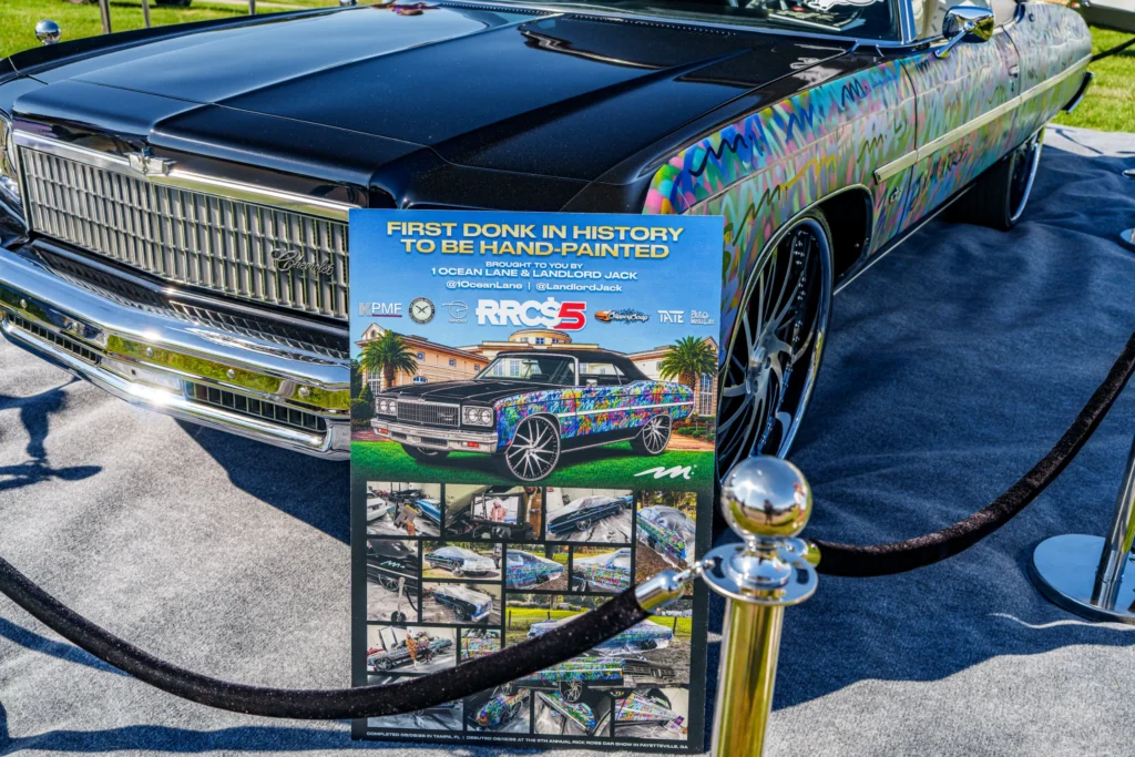 Hand-painted Chevrolet Impala "donk" with black hood and multicolor graffiti-style artwork on sides at outdoor car show