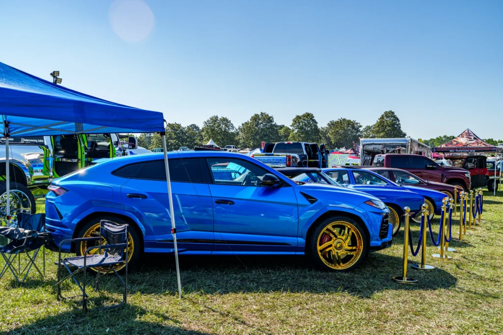 Blue Lamborghini Urus parked beside other vehicles at an outdoor car event with tents and chairs nearby