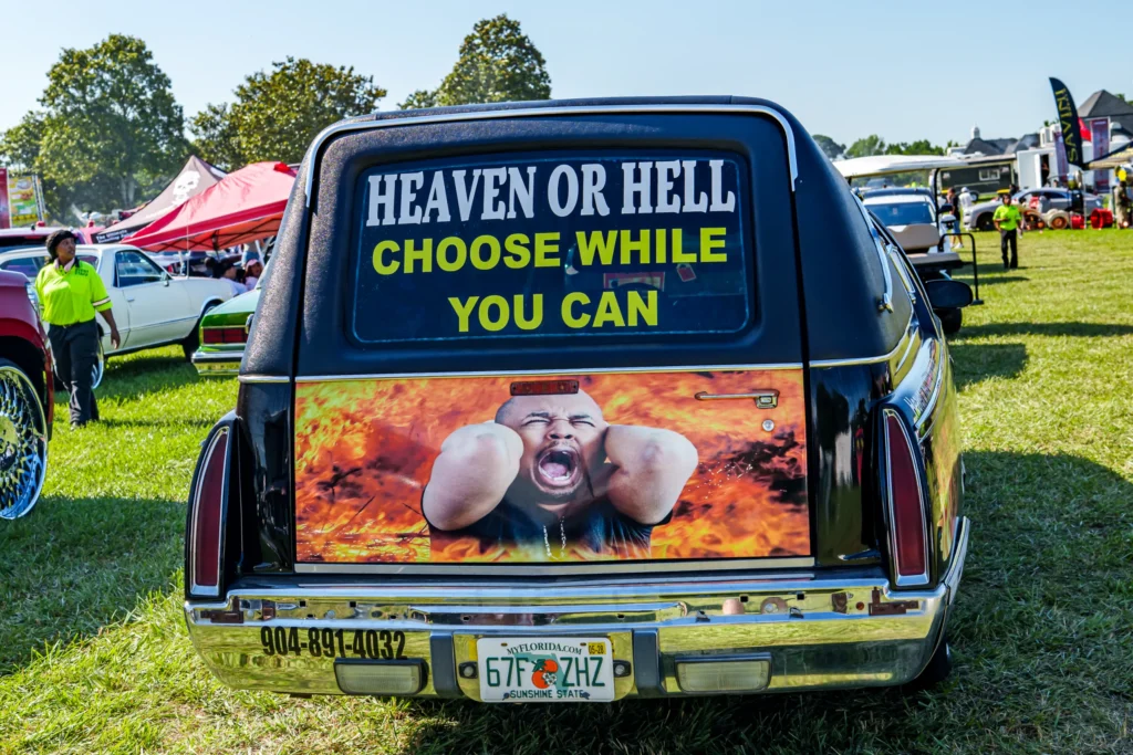 Rear view of black vehicle with custom graphic and 'Heaven or Hell Choose While You Can' decal at outdoor car event