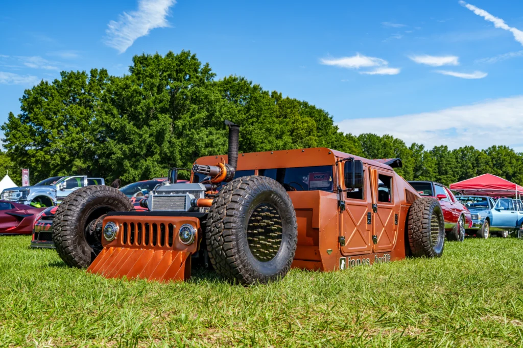 Orange custom low-profile vehicle with large front tires parked on grass at car event with other vehicles nearby