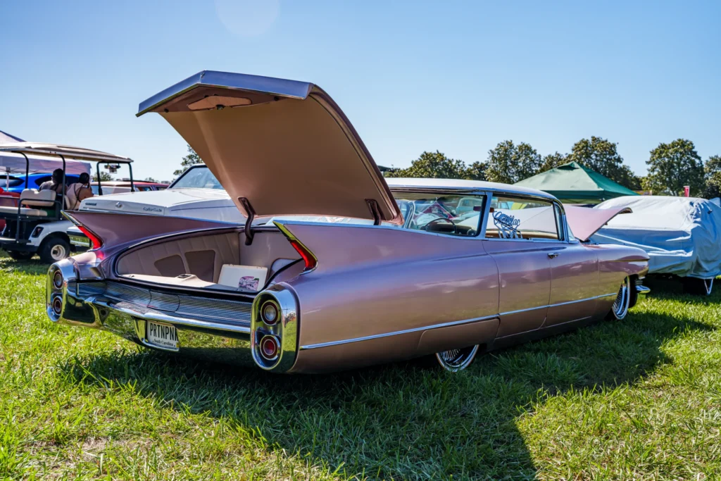 Pink 1959 Cadillac Sedan DeVille with open trunk at an outdoor car event on grass under clear sky