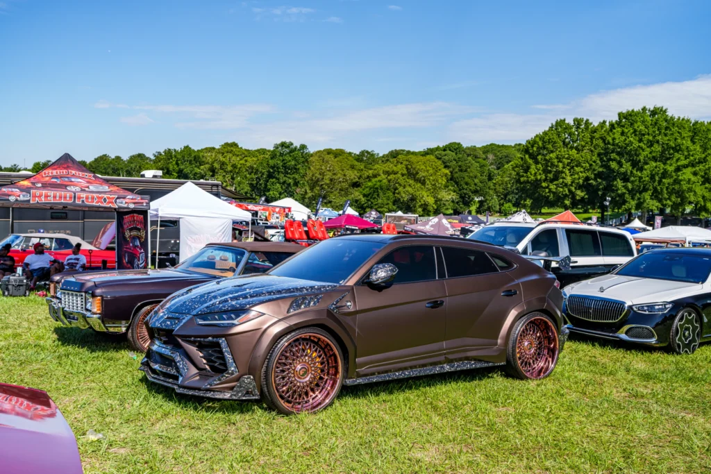 Brown Lamborghini Urus and black Mercedes-Benz SUV parked on grass at outdoor car event with tents and people in background