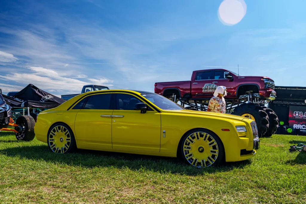 Yellow Rolls-Royce sedan parked on grass with a large red monster truck and tents in background under a blue sky