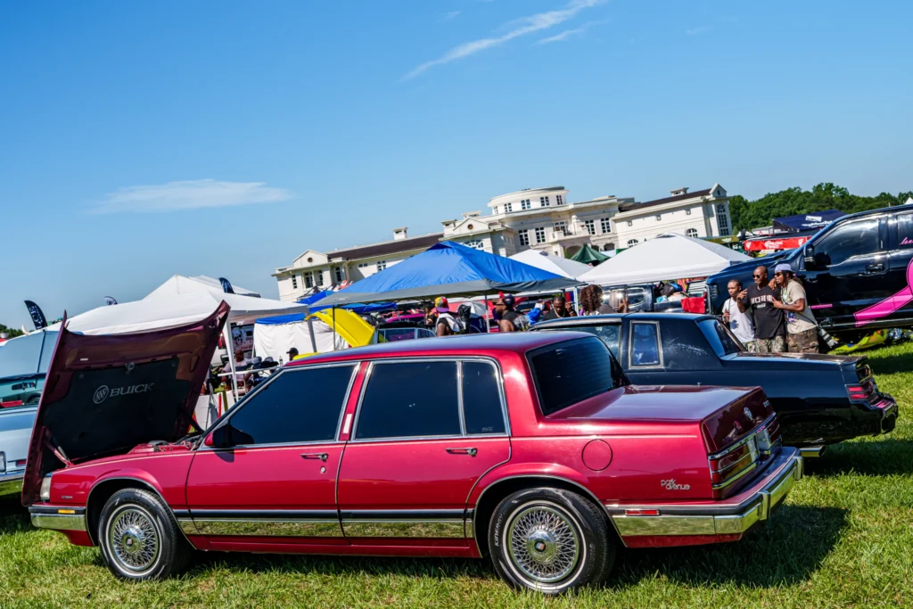 Maroon Cadillac sedan parked on grass at outdoor car event with tents and people in background