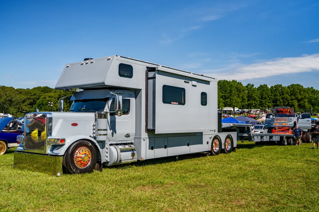 White Peterbilt truck with large camper body, towing a flatbed trailer carrying a lifted blue and orange pickup at an outdoor