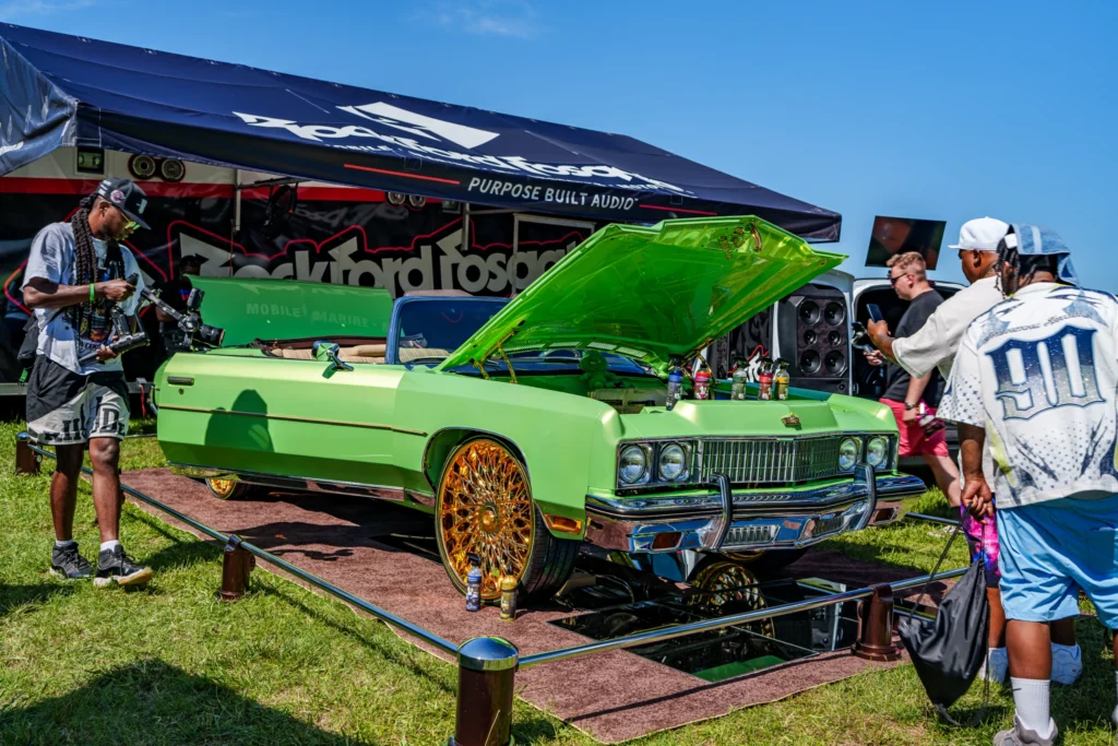 Green convertible Chevrolet Impala with gold wheels and open hood at an outdoor event with people