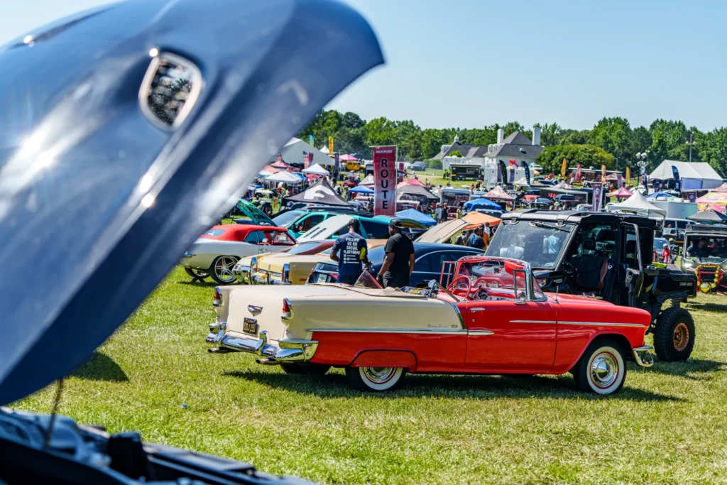 Red and cream Chevrolet Bel Air convertible parked on grass with other vehicles and tents in the background at a car event