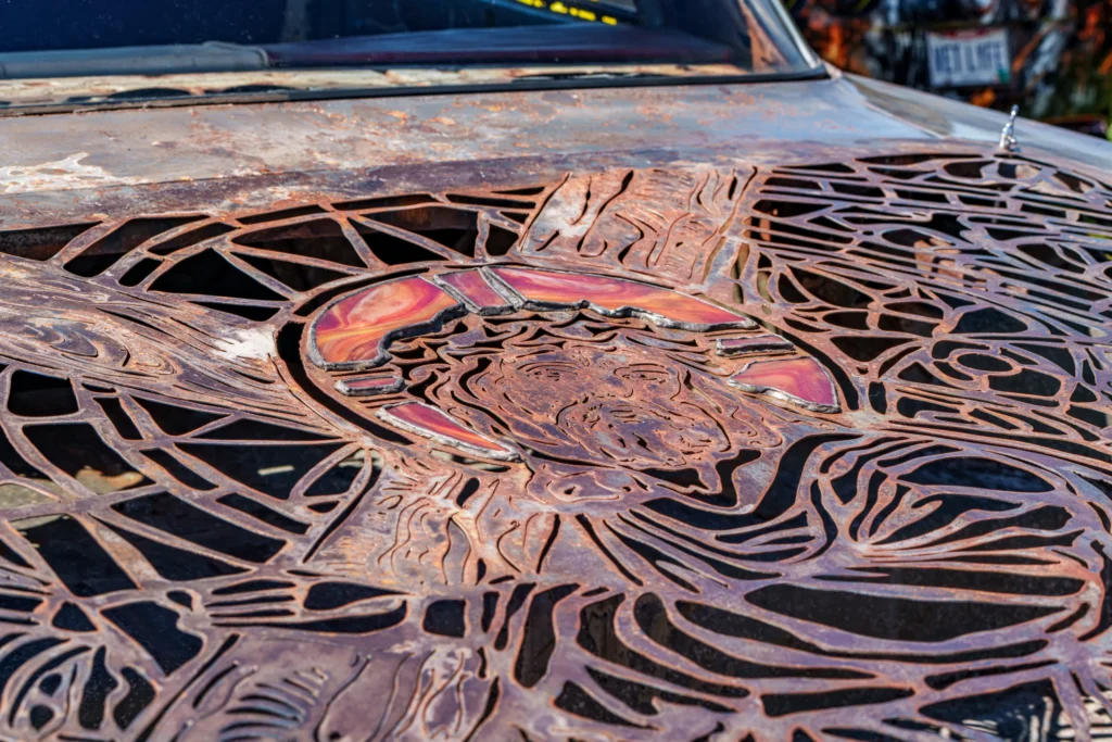 Rusty metal hood with intricate Jesus cutout design and colored inset on a car
