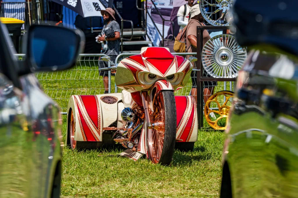 Custom red and white trike motorcycle with detailed paint, on grass at an outdoor event with people and wheels in background
