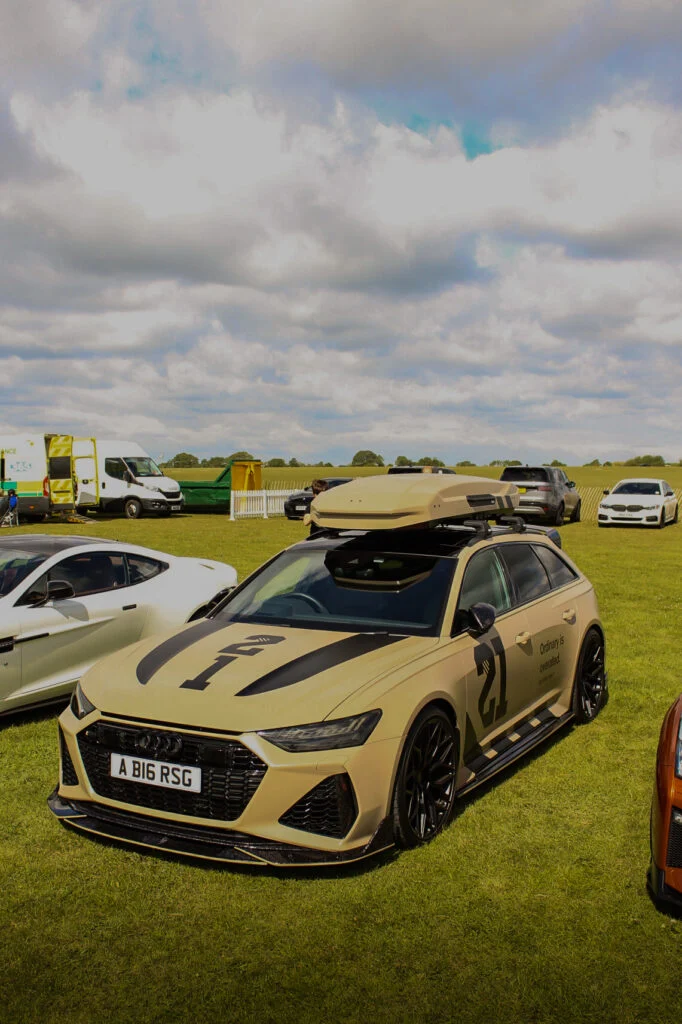 Tan Audi RS6 Avant with roof box on a grassy field at a car event with other vehicles and a partly cloudy sky