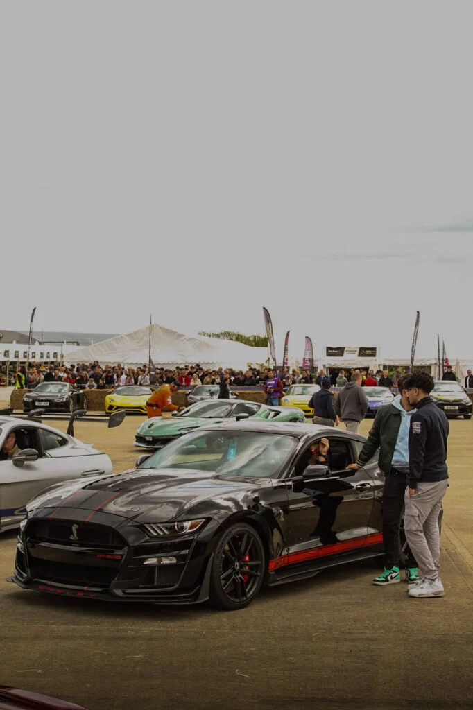 Black Ford Mustang Shelby GT500 parked at an outdoor car event with people and various other cars in the background