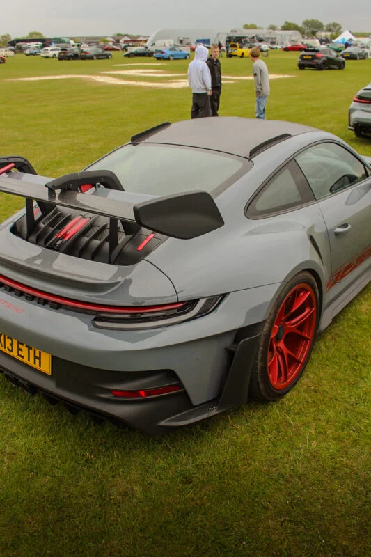 Gray Porsche 911 GT3 RS with red wheels parked on grass at an outdoor car event with people and other vehicles in the backgro