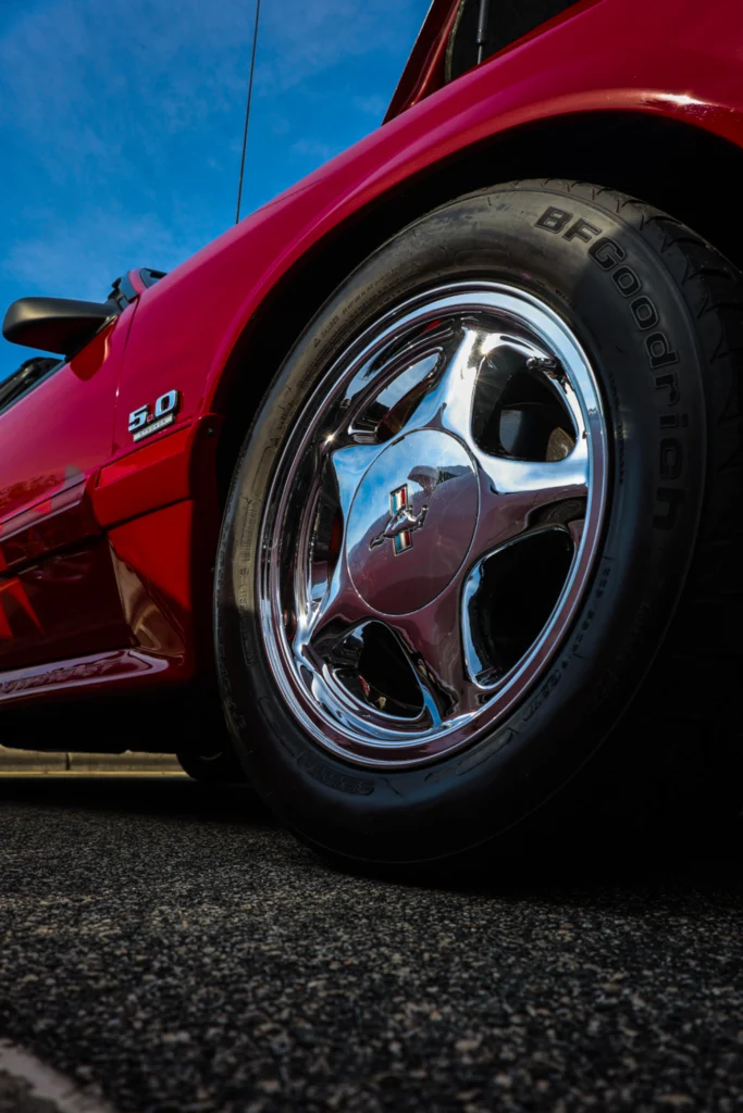 Red Ford Mustang with 5.0 badge and chrome Mustang wheel on asphalt under blue sky