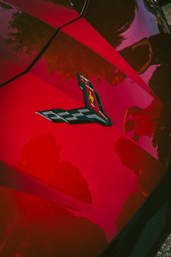 Red Chevrolet Corvette hood with emblem and reflections of people and trees