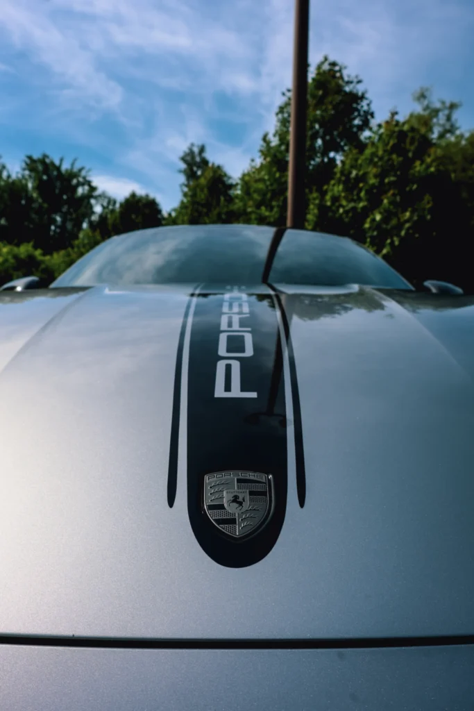 Silver Porsche with black racing stripe on the hood against a background of trees and blue sky