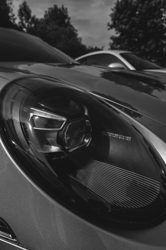 Close view of Porsche headlight and part of a silver coupe parked outside with trees in the background