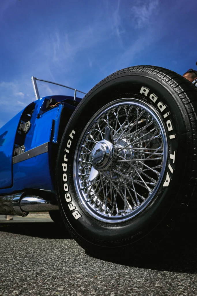 Close-up of wire-spoke wheel and tire on a blue vehicle under clear blue sky at an outdoor event