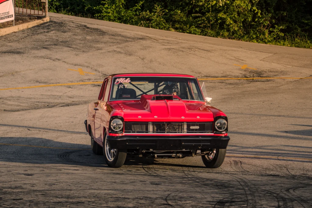 Red 1966 Chevrolet Nova drag car on asphalt track with driver wearing helmet