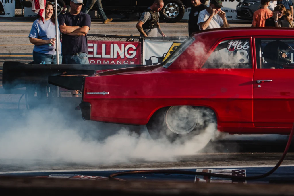 Red 1966 Chevrolet Nova performing a burnout on a drag strip with smoke around rear tires and spectators in background