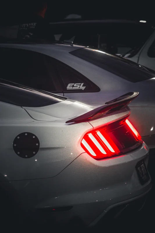 Rear taillight and roof of a white Ford Mustang with a shark fin antenna and ESL sticker on rear side window at night