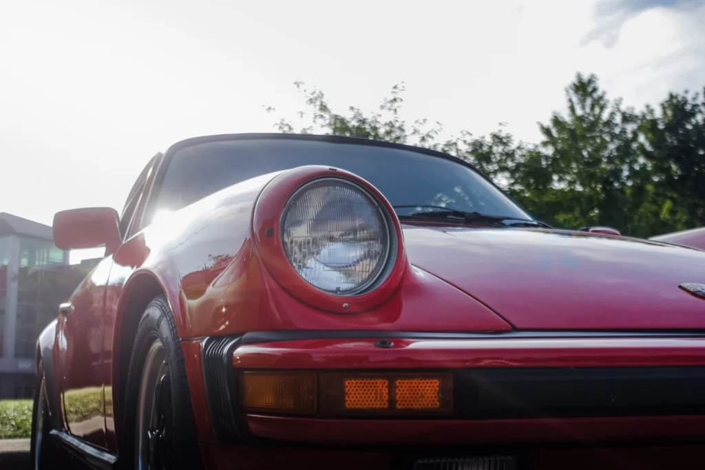 Red Porsche 911 parked outdoors with visible front left headlight and side mirror under daylight