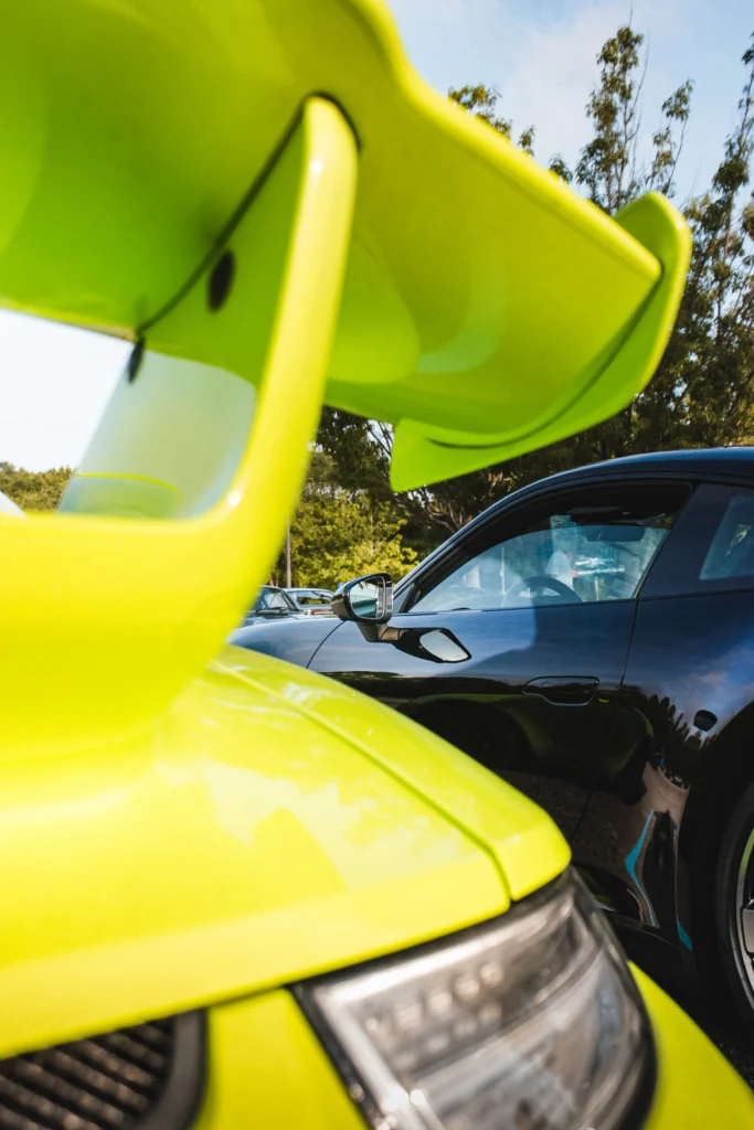 Close-up of a bright yellow Porsche 911 GT3 rear wing and a black coupe parked next to it with trees in the background