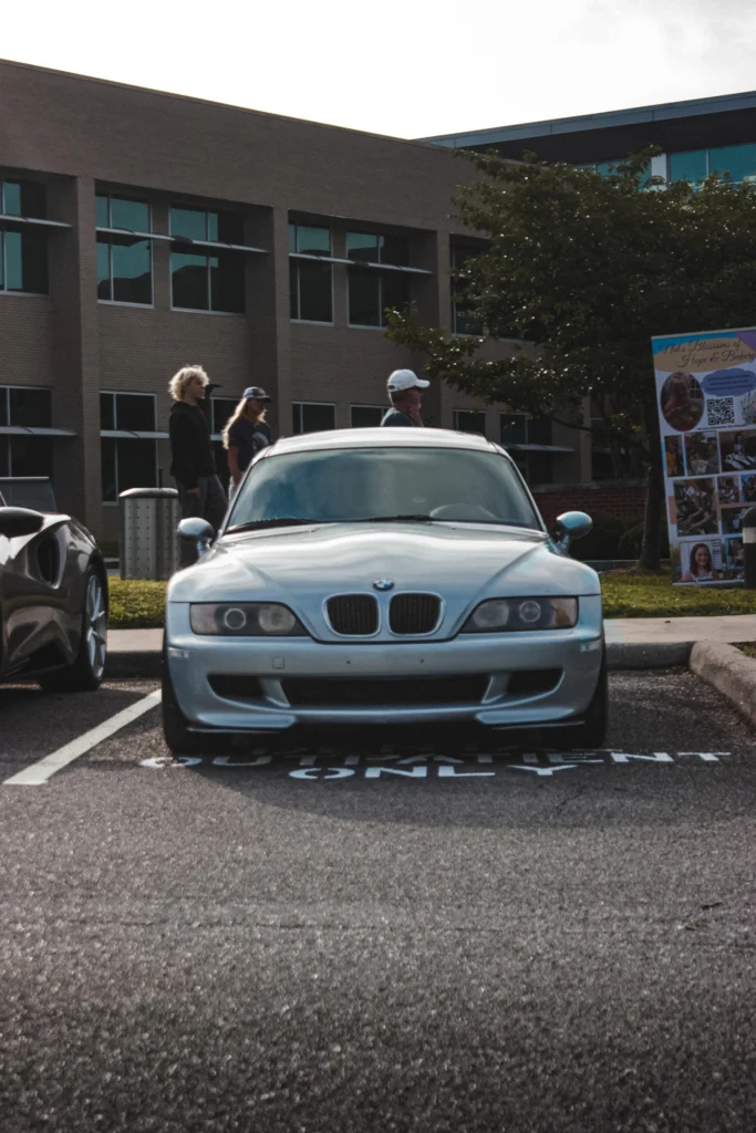 Silver BMW Z3 parked in front of a brick building with three people walking nearby.