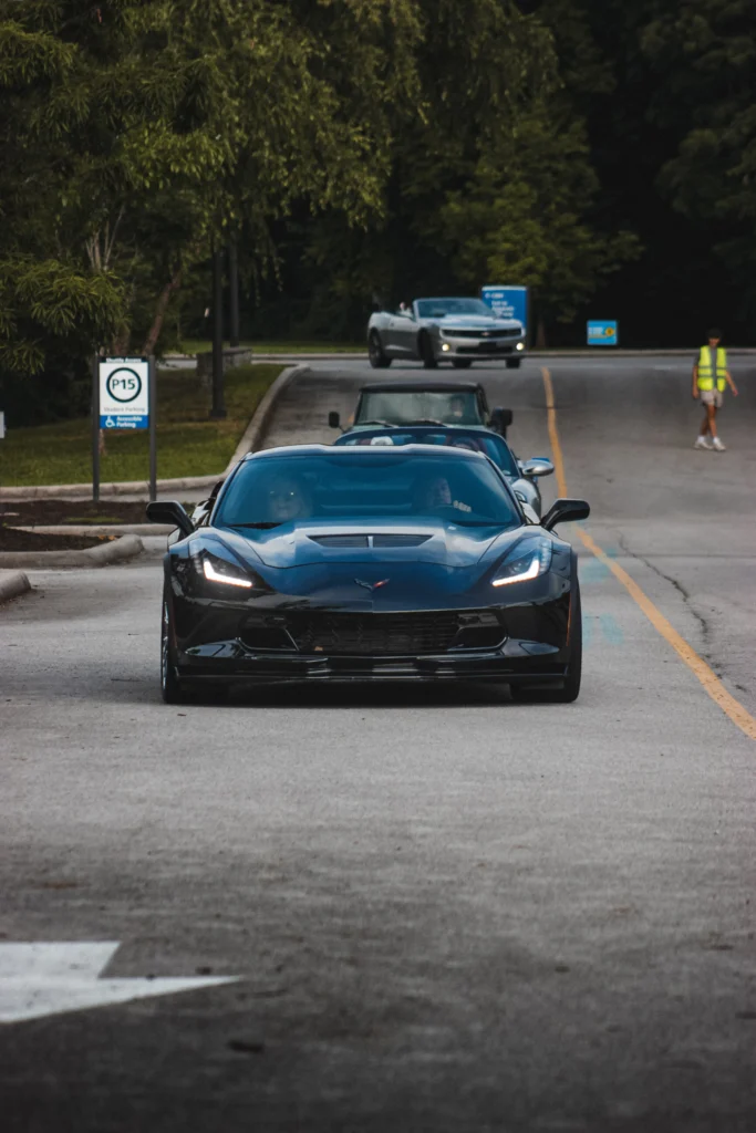 Black Chevrolet Corvette followed by other vehicles on a street near trees and parking signs.