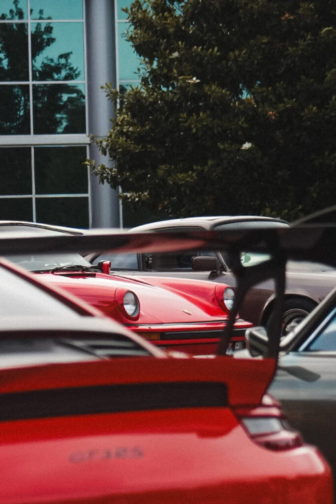 Red Porsche 911 partially visible behind another red Porsche 911 with a large rear wing in an outdoor parking lot