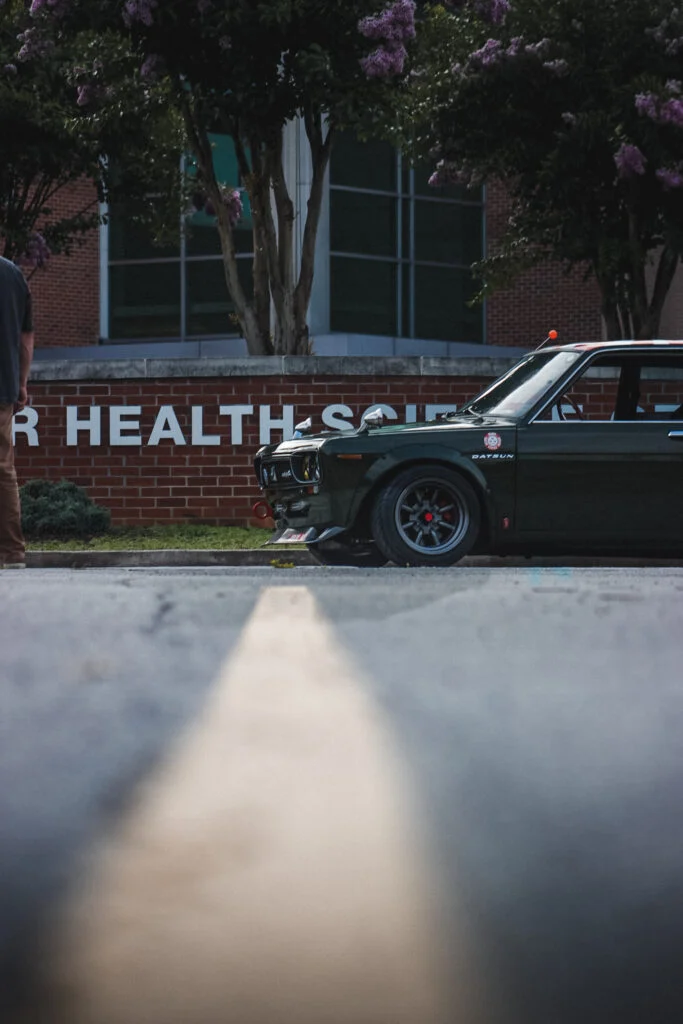 Front 3/4 view of a dark green Datsun coupe parked next to a brick sign with trees in the background