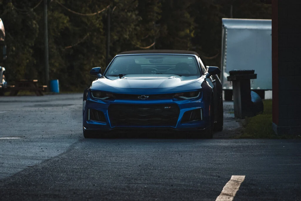 Blue Chevrolet Camaro ZL1 convertible parked on asphalt near a trailer and trees in the background.