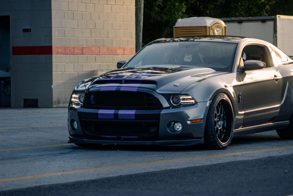 Black Ford Mustang Shelby GT500 with purple racing stripes parked beside a building with a portable toilet in the background