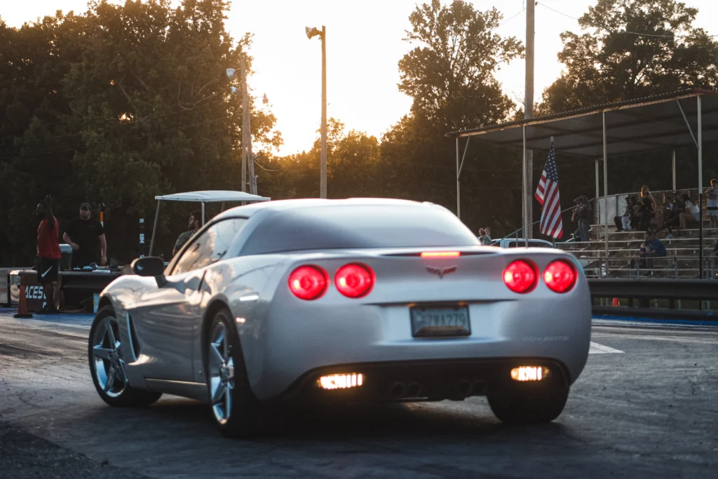Silver Chevrolet Corvette at a drag strip during sunset with people by grandstand and track officials nearby