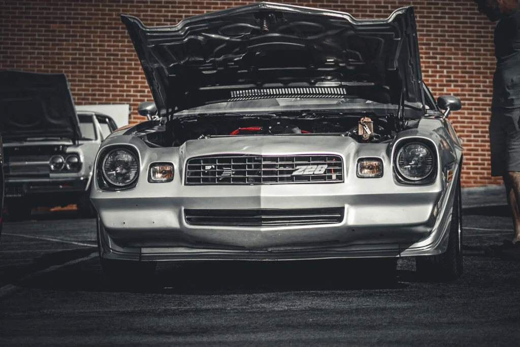 Silver Chevrolet Camaro Z28 with hood open parked outdoors near a brick wall, with a white car partially visible in backgroun