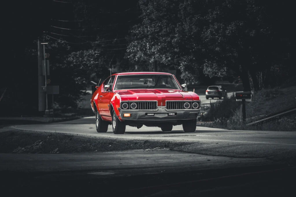 Red Oldsmobile Cutlass driving on a rural road with trees and other vehicles in the background