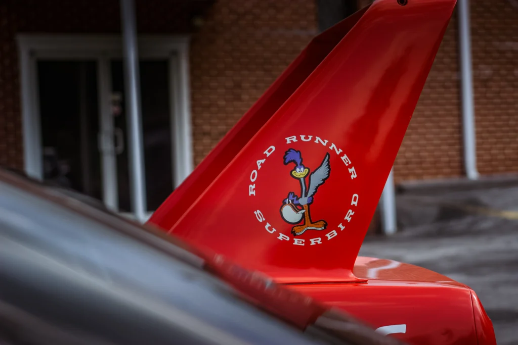 Red Road Runner Superbird rear wing with Road Runner cartoon and text, with blurred cars and brick building in background