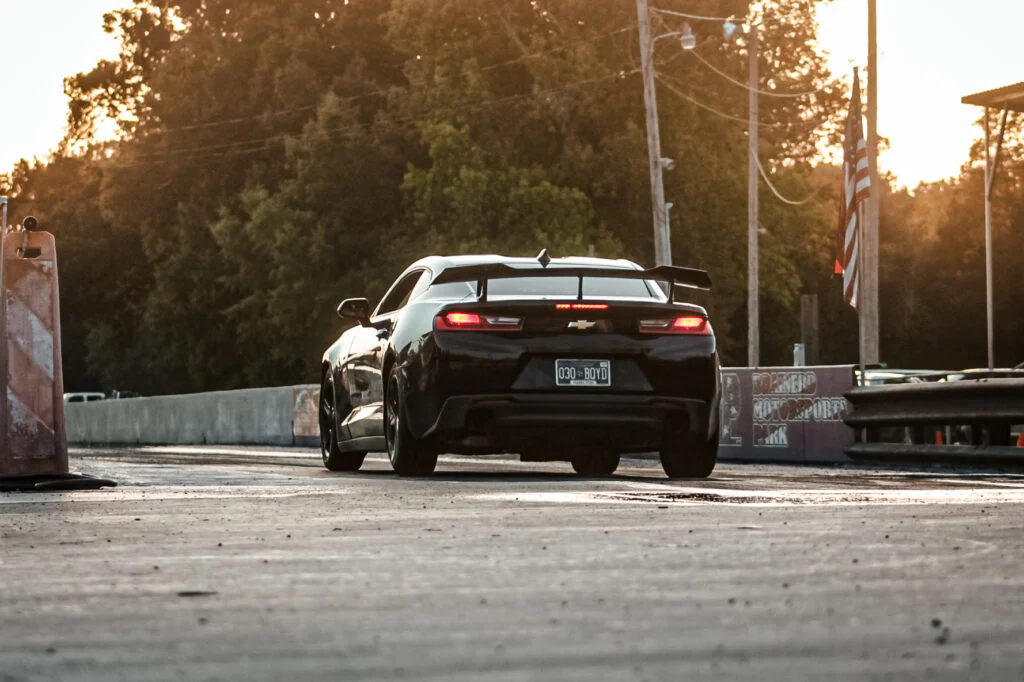 Black Chevrolet Camaro with rear spoiler on a drag strip at sunset with trees and poles in the background