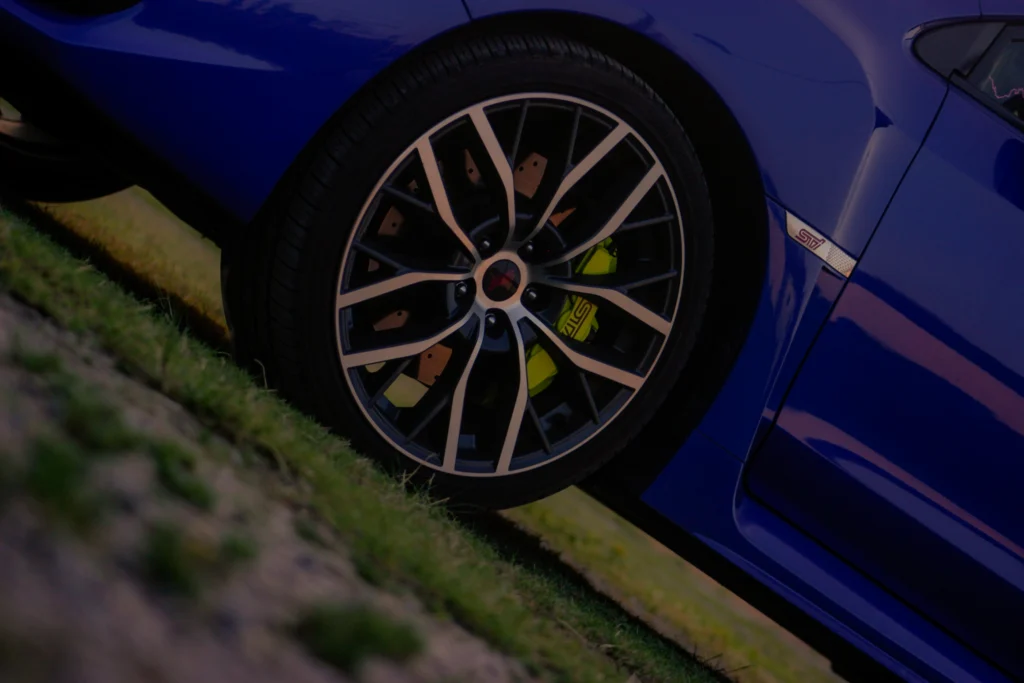 Close-up of a blue Subaru WRX STI front wheel and brake caliper with STI logo visible, parked on grass.