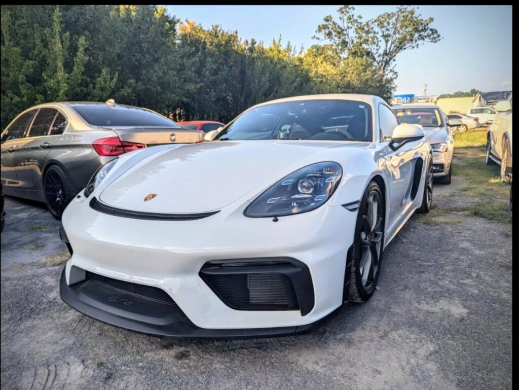 White Porsche 718 Cayman GT4 parked in an outdoor lot with other cars and trees in the background
