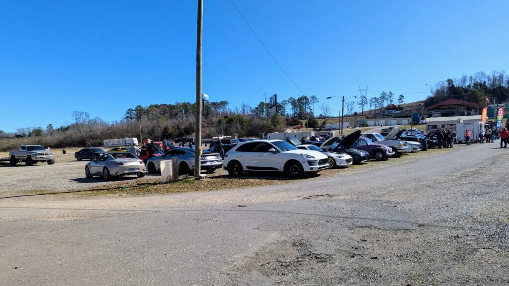 Lineup of various cars including white Porsche Macan, white Mazda MX-5, and purple Volkswagen Beetle at an outdoor car meet o