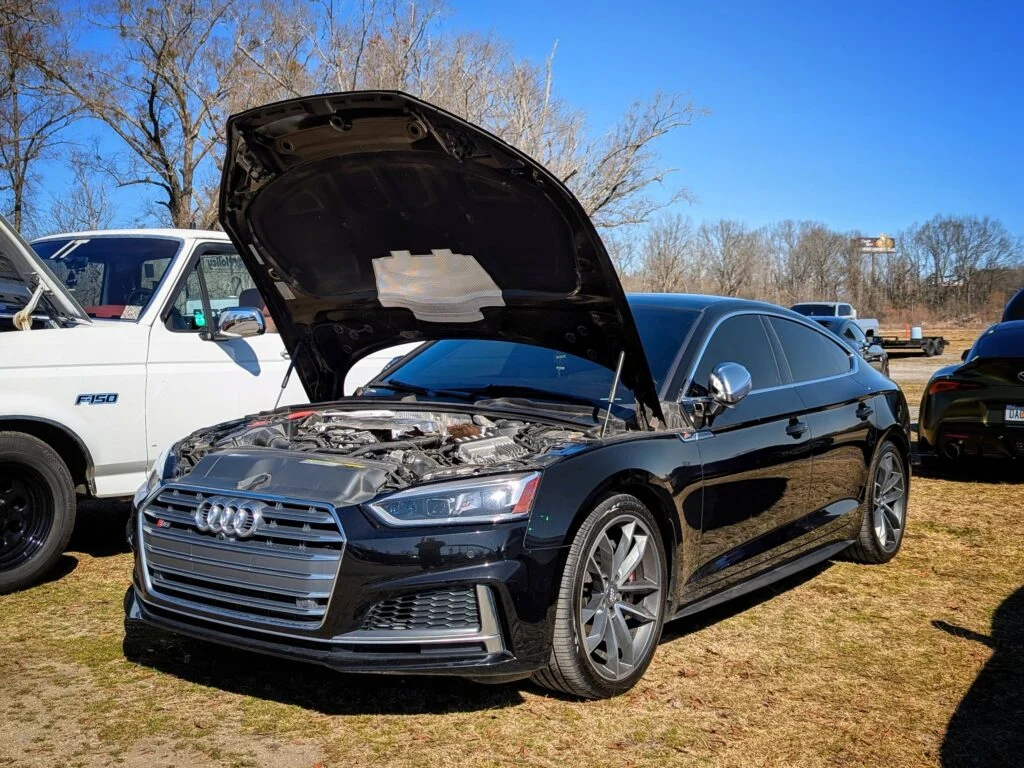 Black Audi S5 with hood open at outdoor car event on grass with other vehicles around under clear blue sky