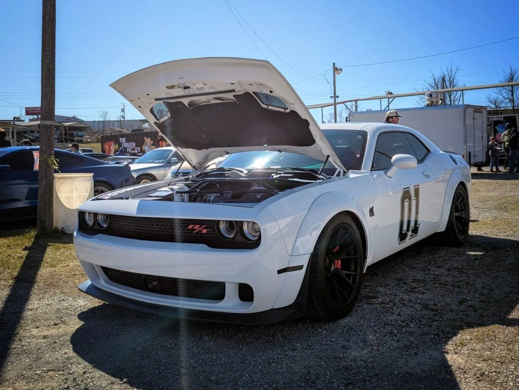 White Dodge Challenger R/T with raised hood parked in a gravel lot at a car event on a clear day.
