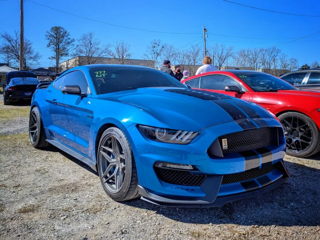 Blue Ford Mustang Shelby GT500 and red Ford Mustang parked in a gravel lot with people and other cars in background