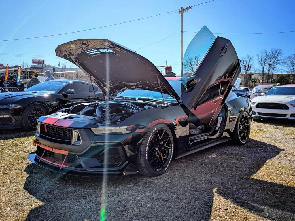 Black Chevrolet Camaro with hood and doors open at an outdoor car show with other vehicles and people in background
