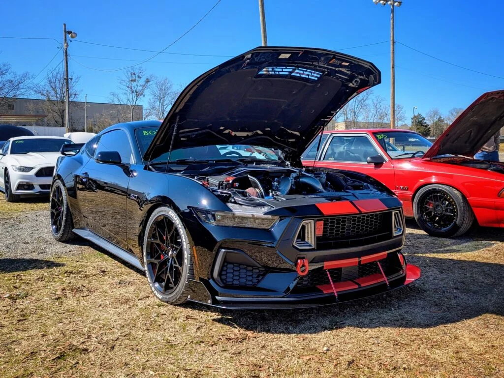 Black Ford Mustang with hood open revealing engine at an outdoor car meet with other parked cars nearby